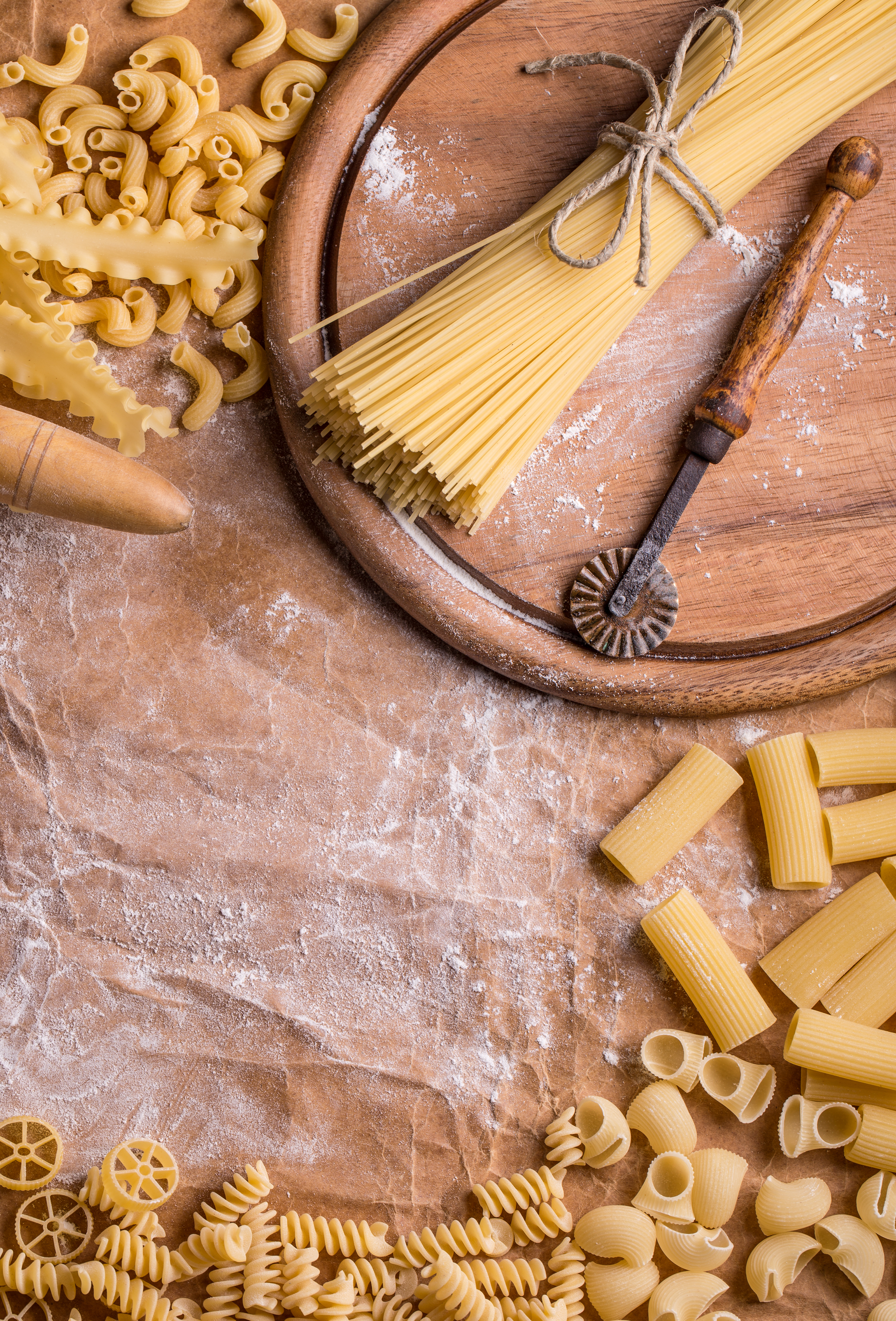 Various uncooked pasta types and a wooden rolling pin on a floured surface.