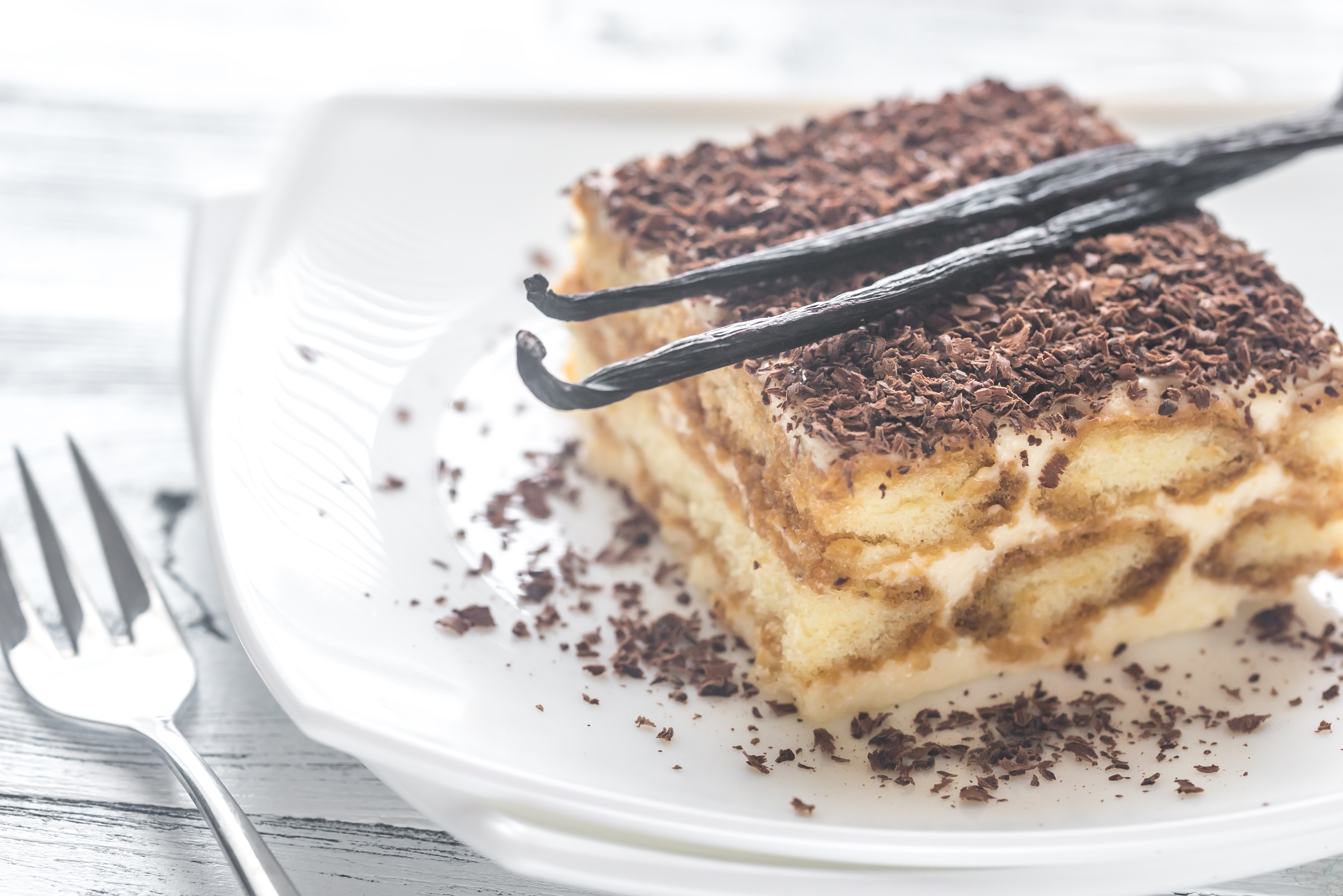 A delicious slice of tiramisu with chocolate shavings and vanilla pods on a white plate.