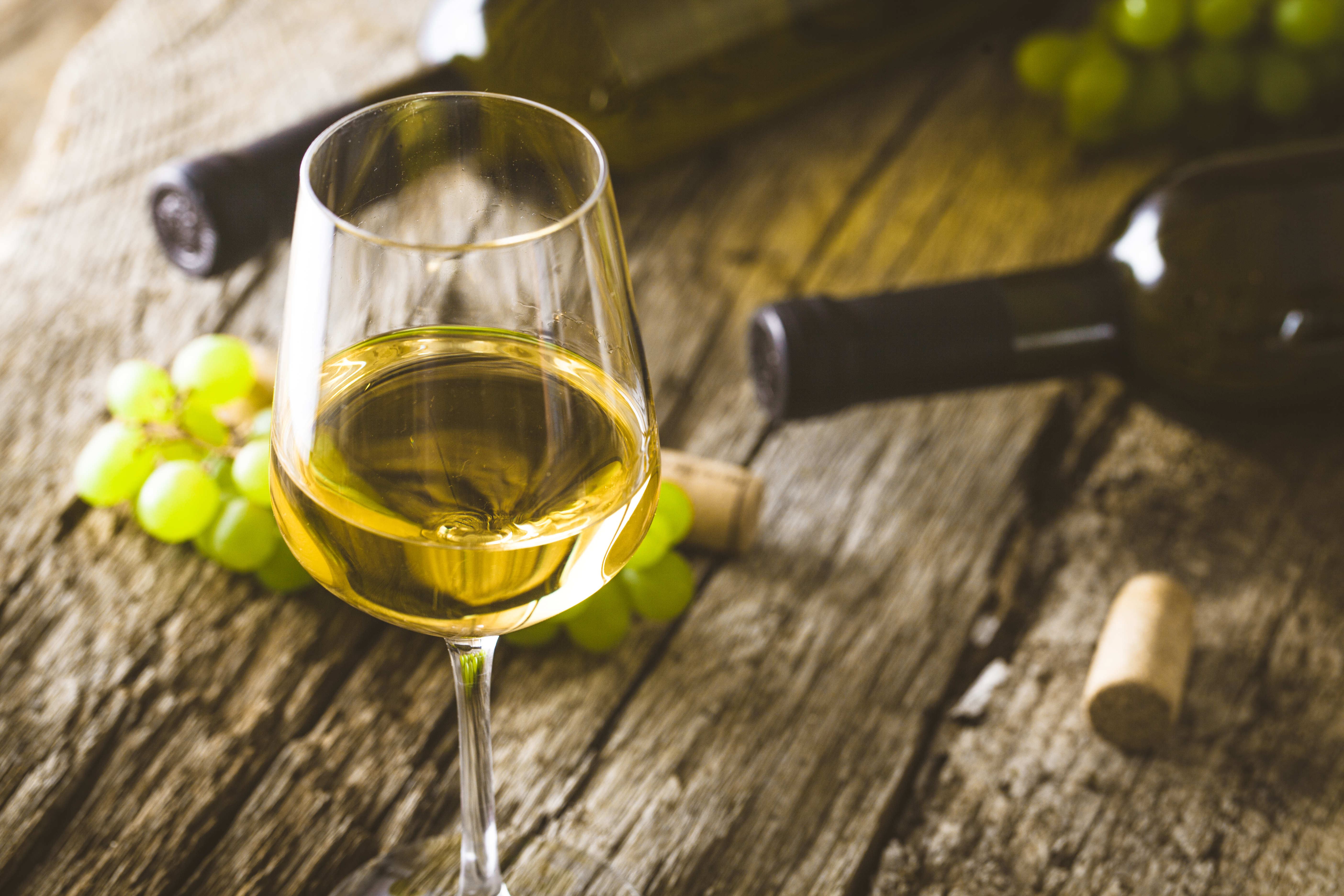 A glass of white wine with grapes and wine bottles on a rustic wooden surface.