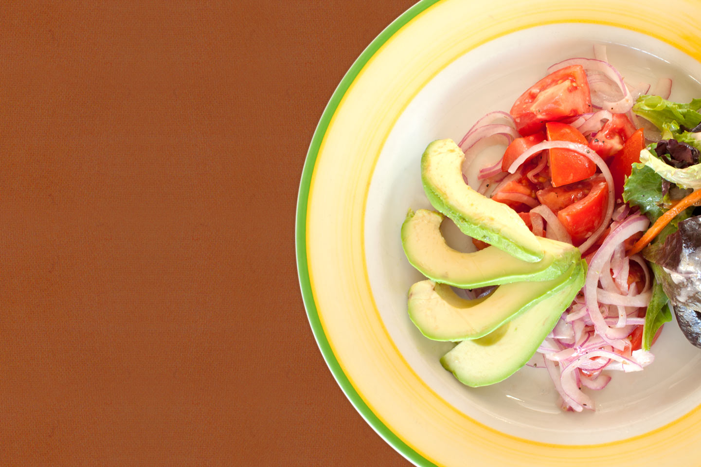 Fresh avocado, tomato, and salad on a plate with a brown background.